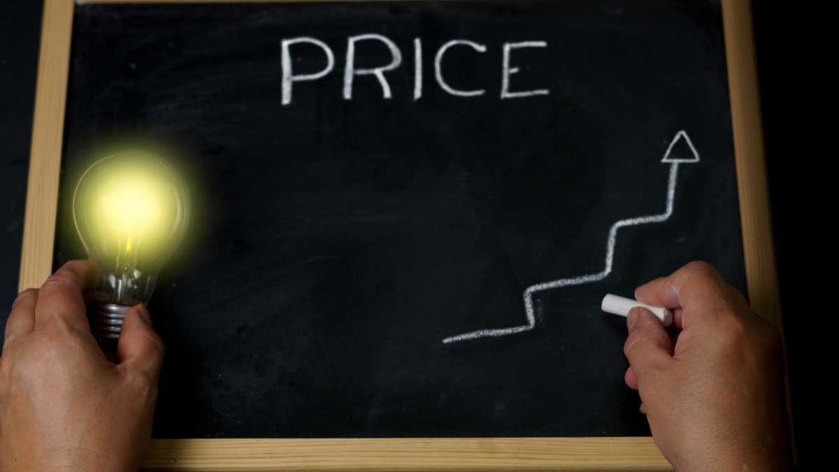 Hand holding a lit bulb and drawing an uptrend graph labelled PRICE on a blackboard