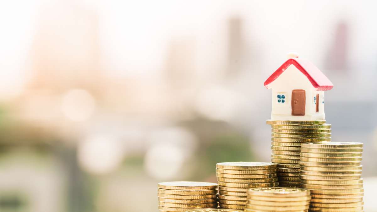 a stack of gold coins with a small house model on top, against a blurred cityscape background