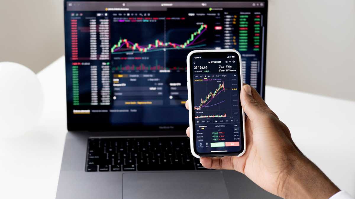 A persons hand holding a smartphone with stock market data in front of a laptop showing trading charts