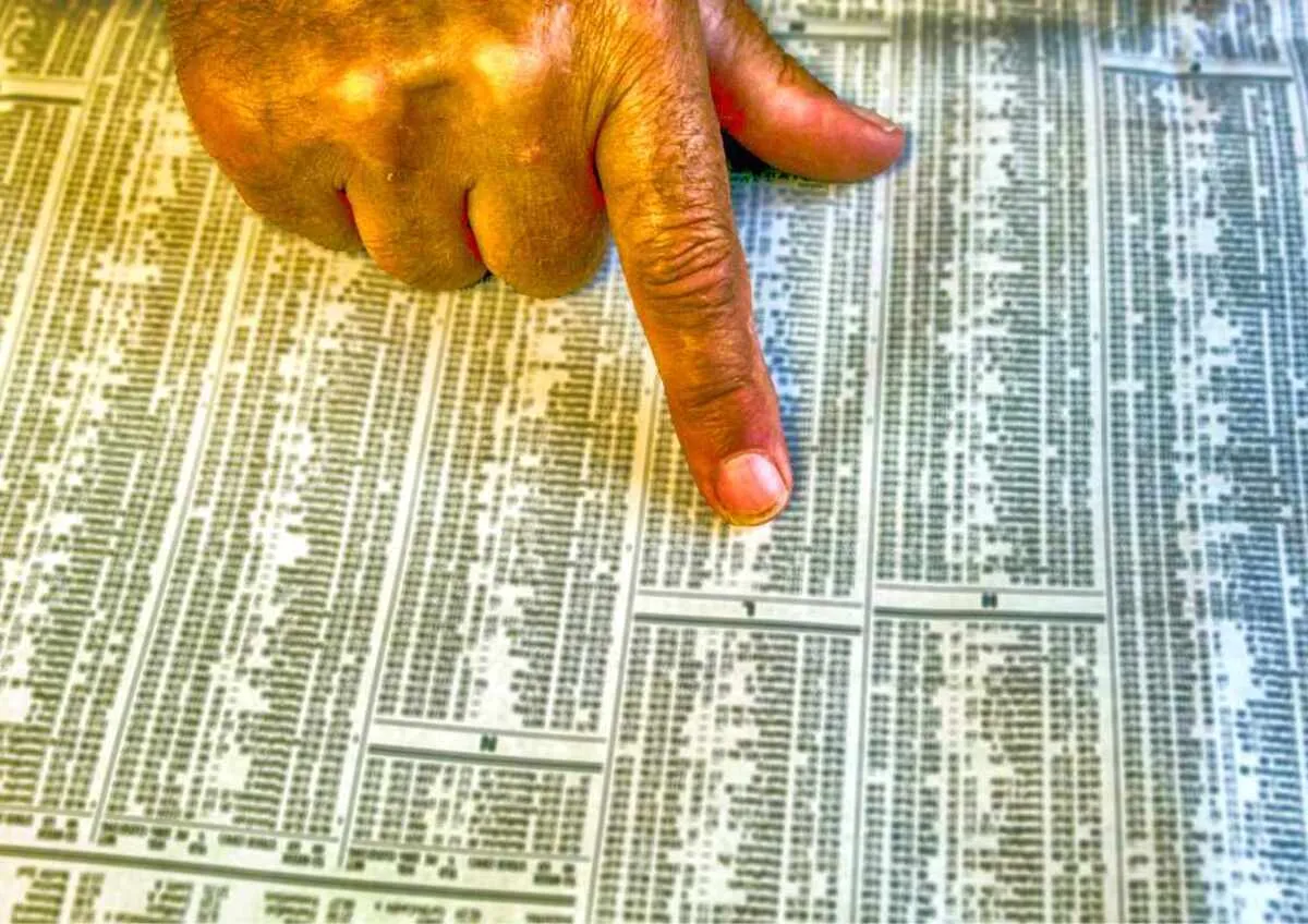 Hand pointing at stock market listings in a newspaper.