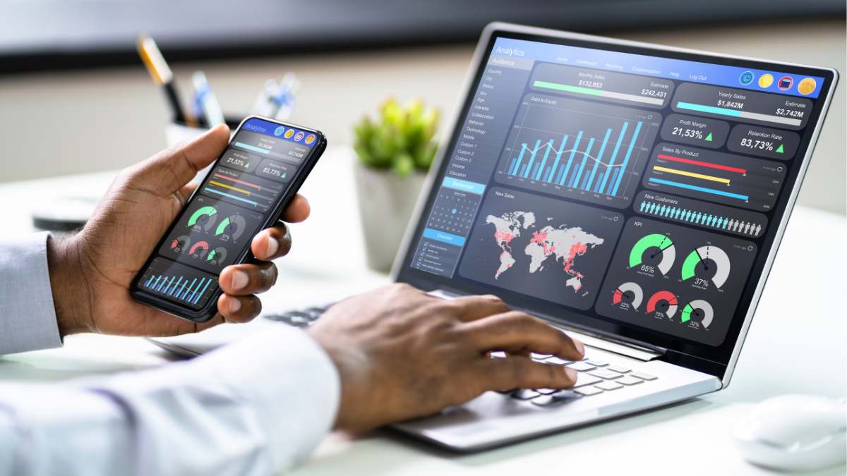 a person's hands interacting with both a smartphone and a laptop, which display colourful graphs and analytics dashboards.