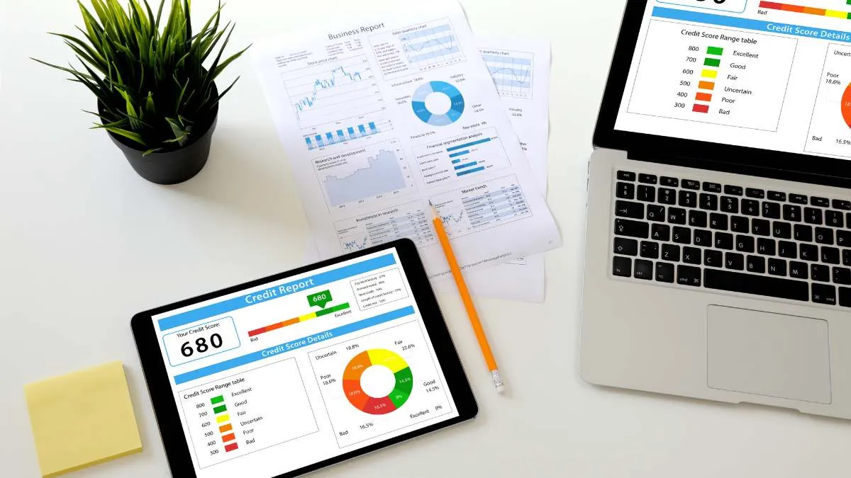 a workspace with a tablet displaying a credit score report, alongside a laptop showing various business reports and graphs.