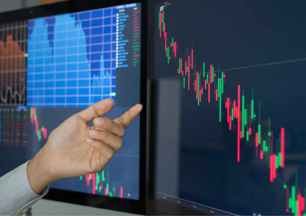 Hand pointing at financial graphs on computer screens.