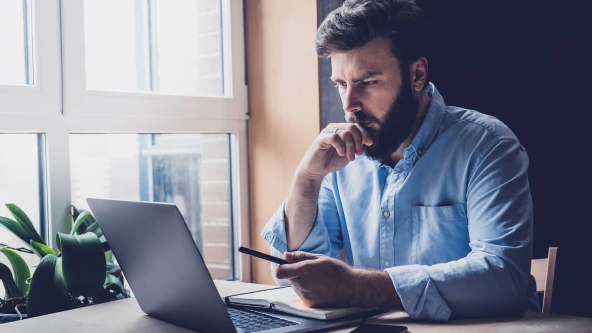 A bearded individual engaged in thought pen in hand in front of a laptop screen