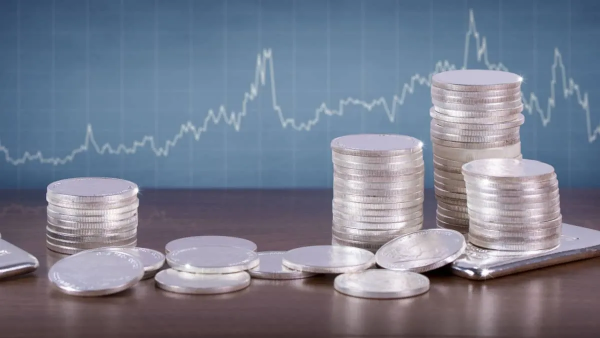 Silver bars and coins on a wooden table with a price chart at the back