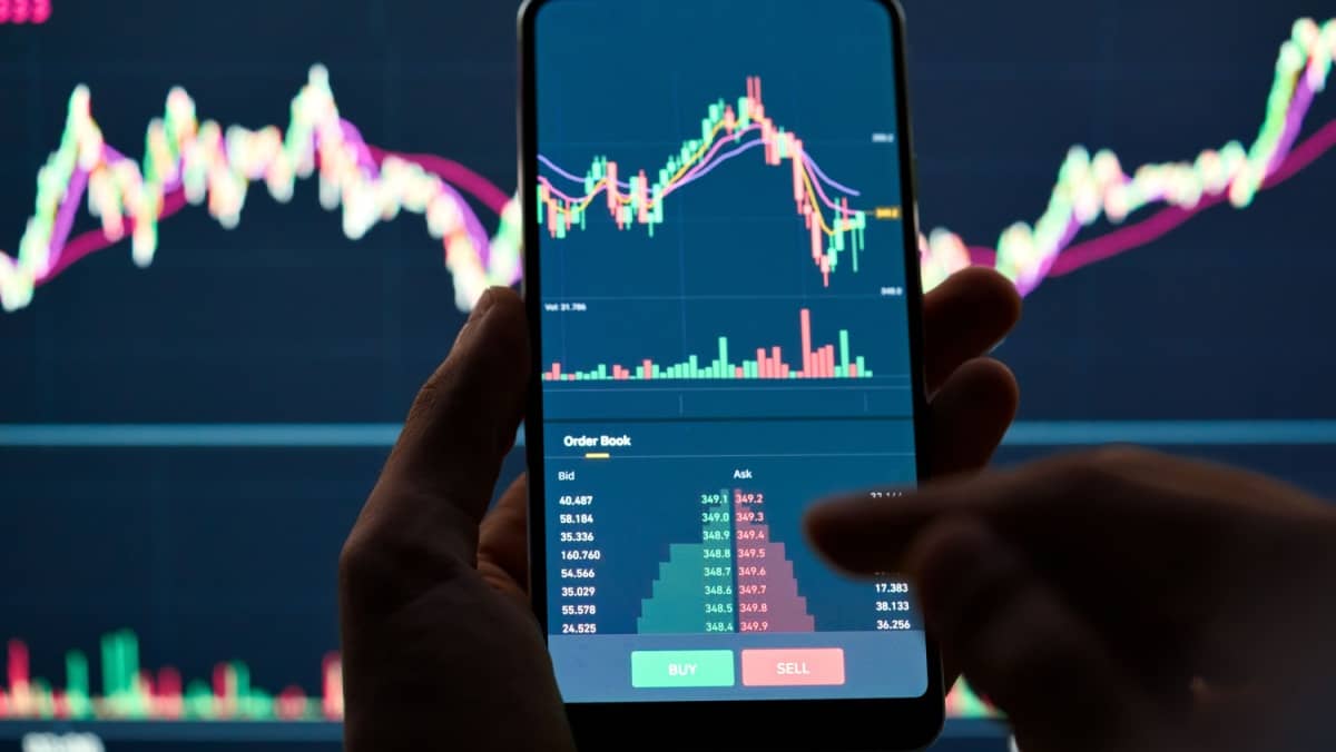 Someone is using a mobile device while trading online with a background showing a big monitor screen of a trading platform