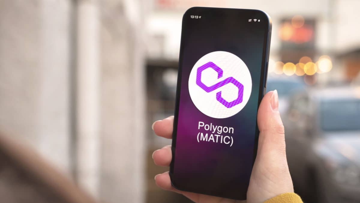 A hand holding a smartphone, with the screen displaying the Polygon crypto icon