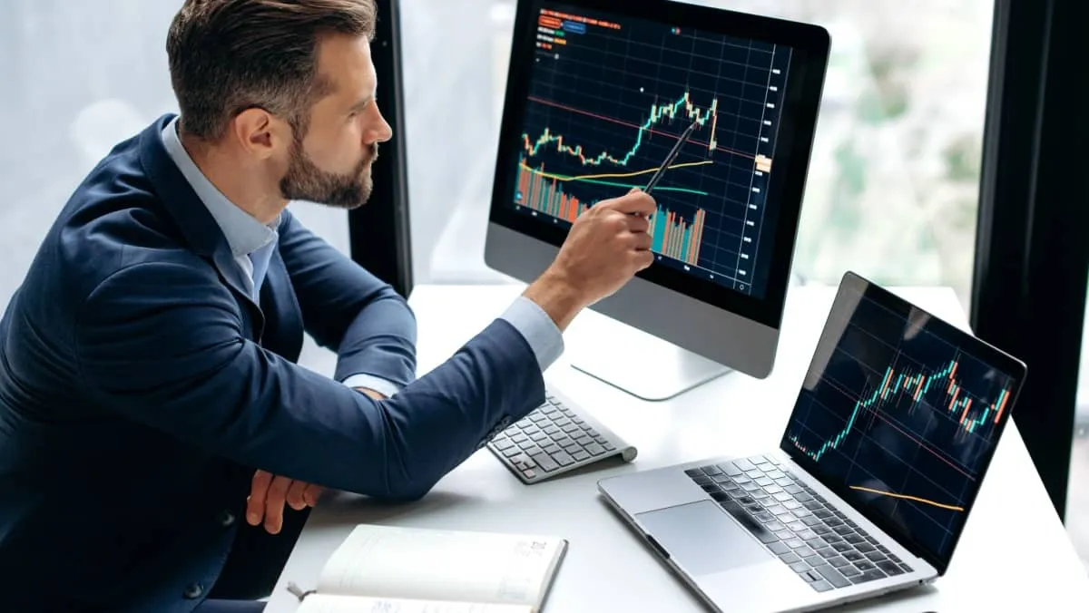 A man gesturing with a pen drawing attention to a trading screen on the left accompanied by a laptop on the right