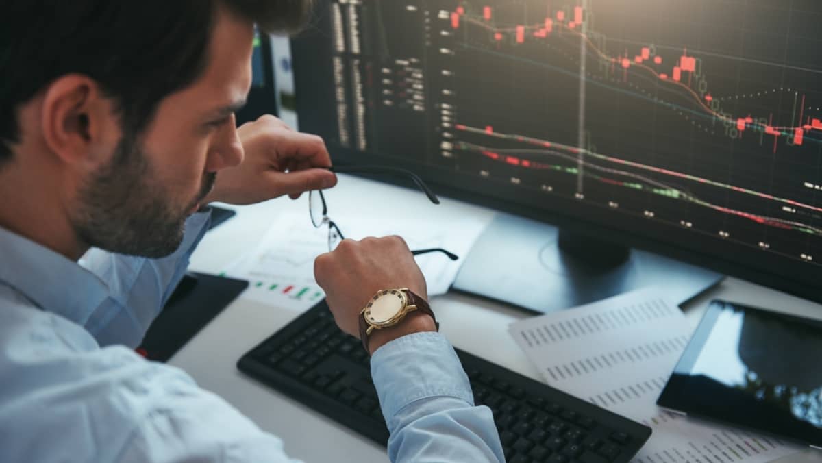 A male trader glancing at his watch while actively engaging with data and charts on a computer screen