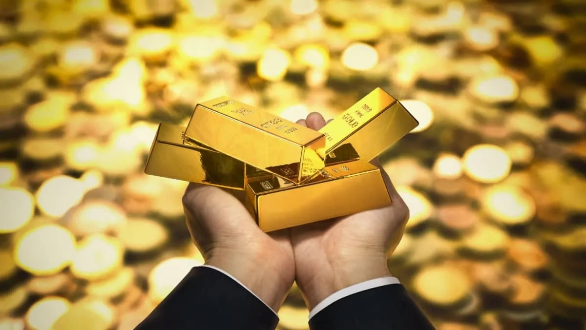 Hands showing gold bars against the backdrop of a blurred sea of gold coins below