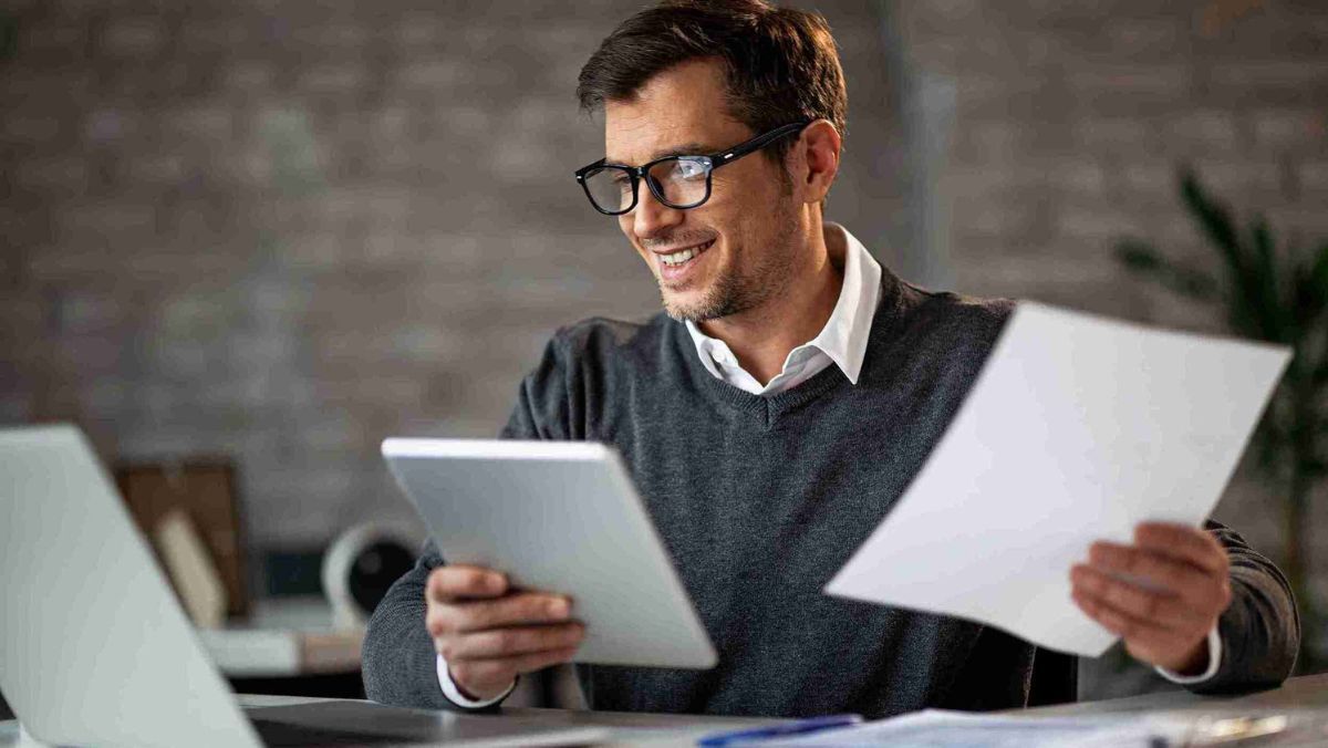 Smiling man at work holding documents and looking at laptop screen