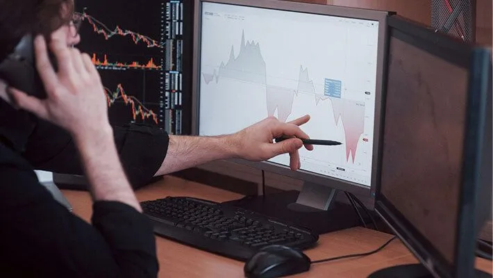 A trader on a phone call gestures towards the computer screen displaying a trading chart