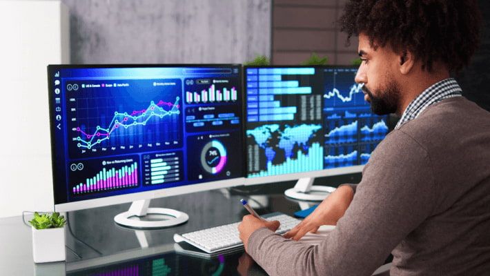  A man sits at a desk with multiple screens displaying data charts, taking notes.