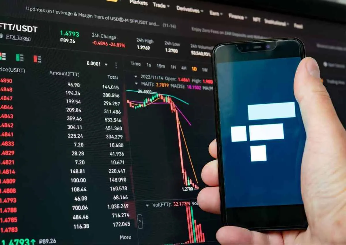 A hand holding a smartphone displaying a financial trading app in front of a stock market screen.