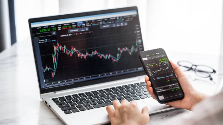 A person trading financial chart on mobile with computer screens in the background